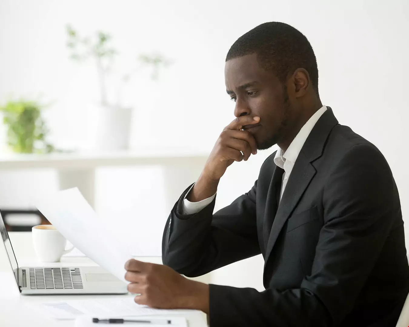En man i mycket prydliga kontorskläder, kostym, sitter vid dator. Håller i ett papper, ser mycket bekymrad ut och har en hand framför munnen i en djupt fokuserad gest.
