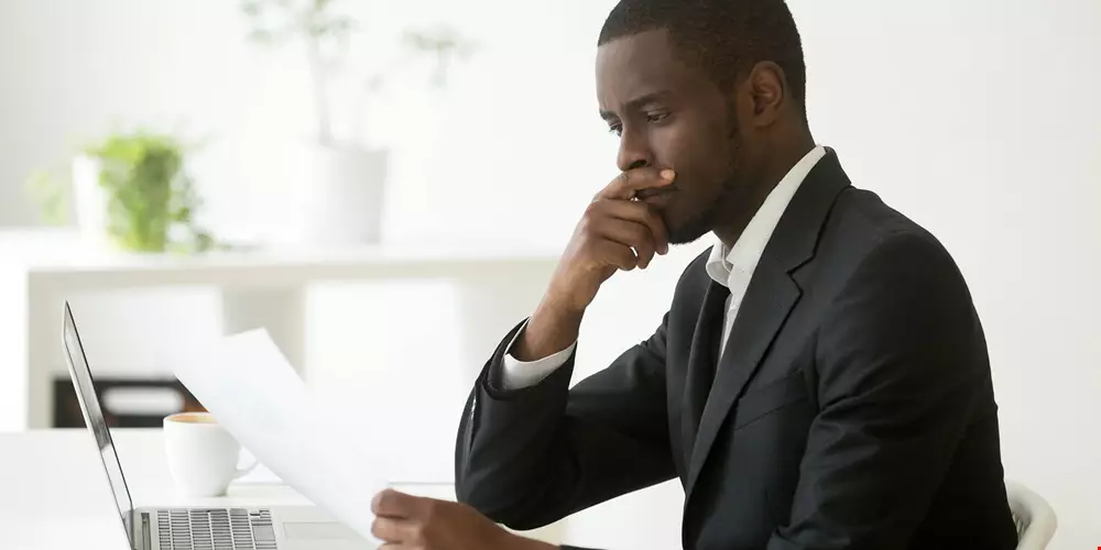 En man i mycket prydliga kontorskläder, kostym, sitter vid dator. Håller i ett papper, ser mycket bekymrad ut och har en hand framför munnen i en djupt fokuserad gest.