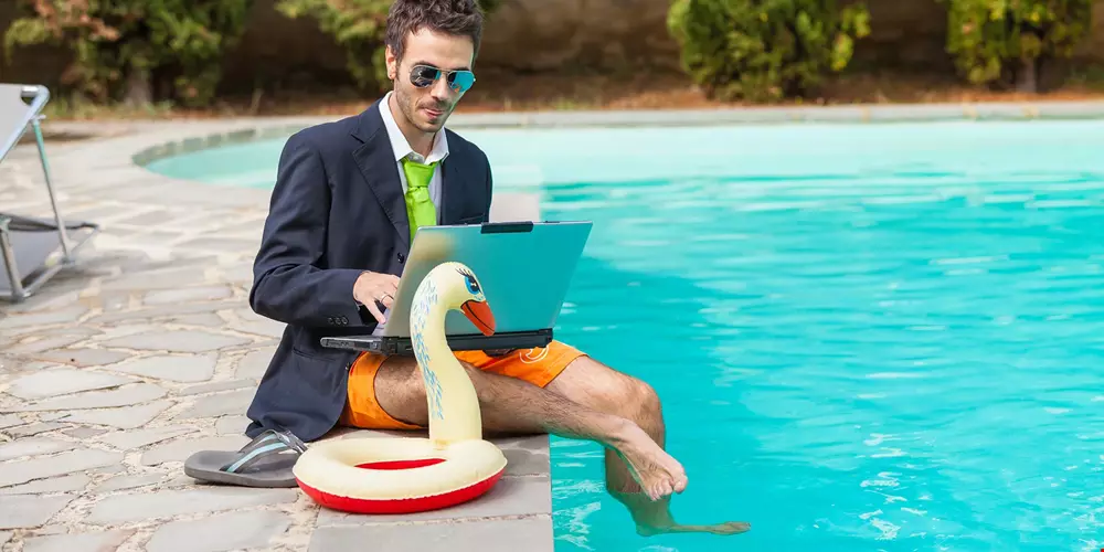 En man i kostym sitter med dator i knäet vid en pool, med fötterna ner i vattnet. Ser ut som ledighet, men jobbar