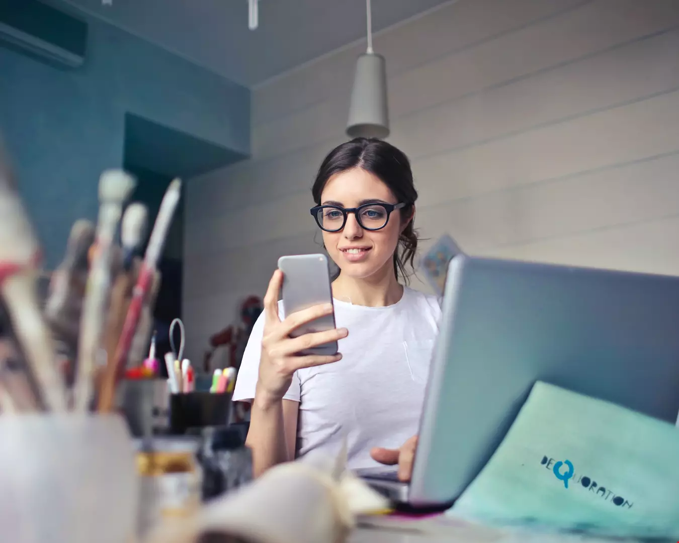 Ung kvinna jobbar framför datorn, sitter med mobilen i handen, kvinnan har glasögon med svart båge.