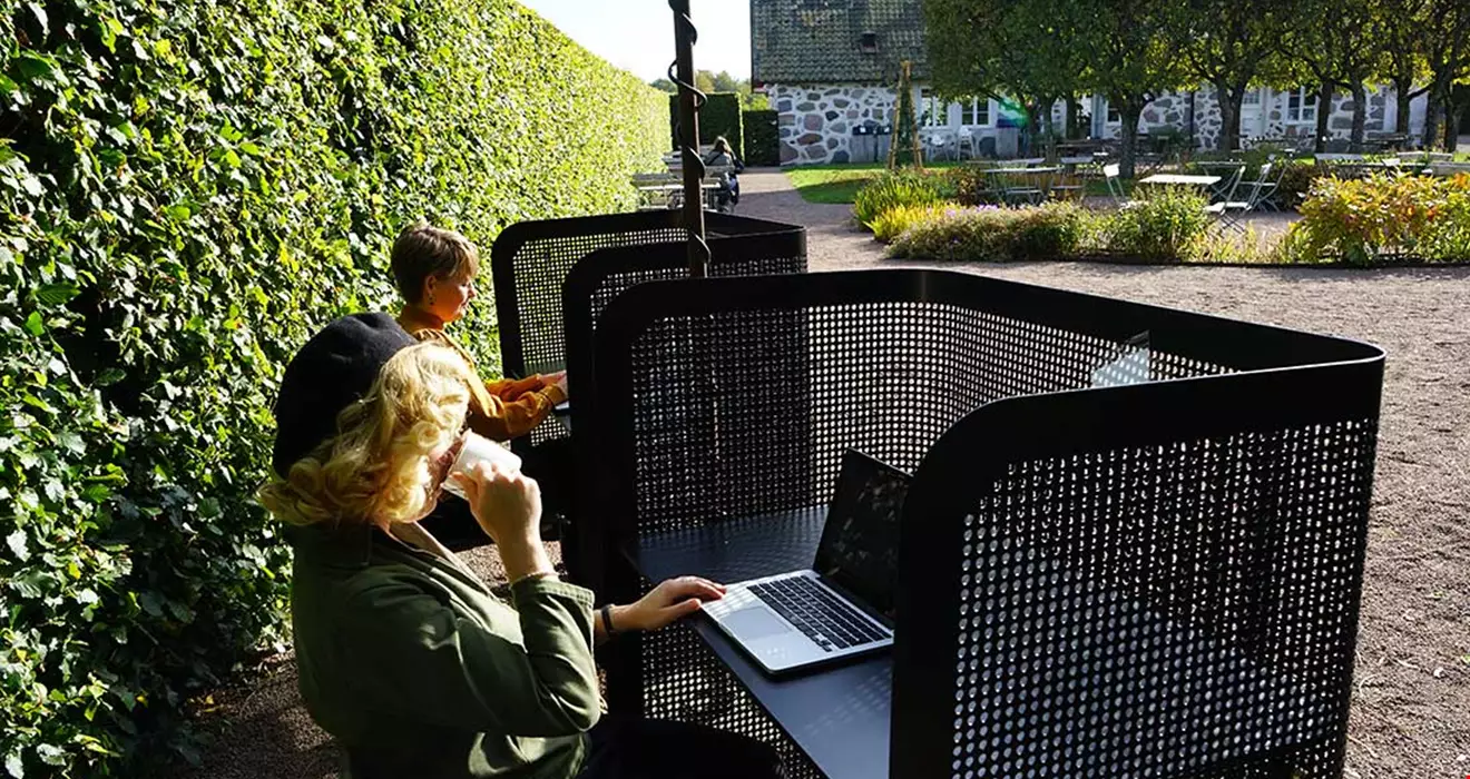 Två personer sitter ute i en park vid varsitt skrivbord. Skrivbordet har solskydd, det står en bärbar dator på vardera bord.