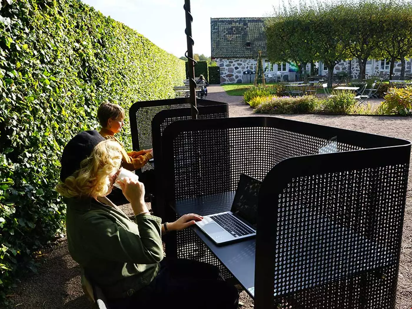 Två personer sitter ute i en park vid varsitt skrivbord. Skrivbordet har solskydd, det står en bärbar dator på vardera bord.