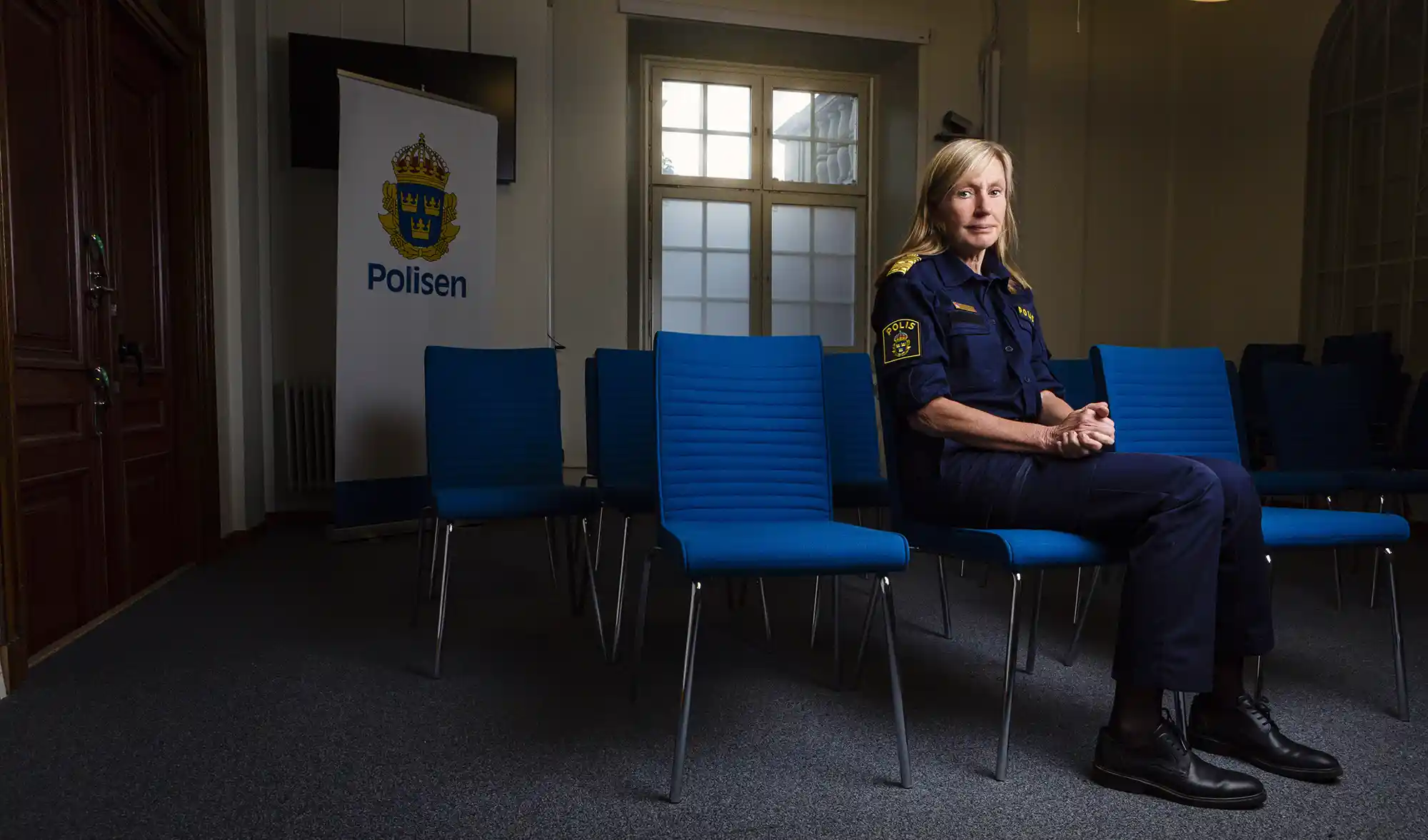 Blond kvinna med pagefrisyr, övre medelåldern, sitter ner i ett rum fullt av tomma stolar. Hon är klädd i polisuniform och ser allvarlig ut.