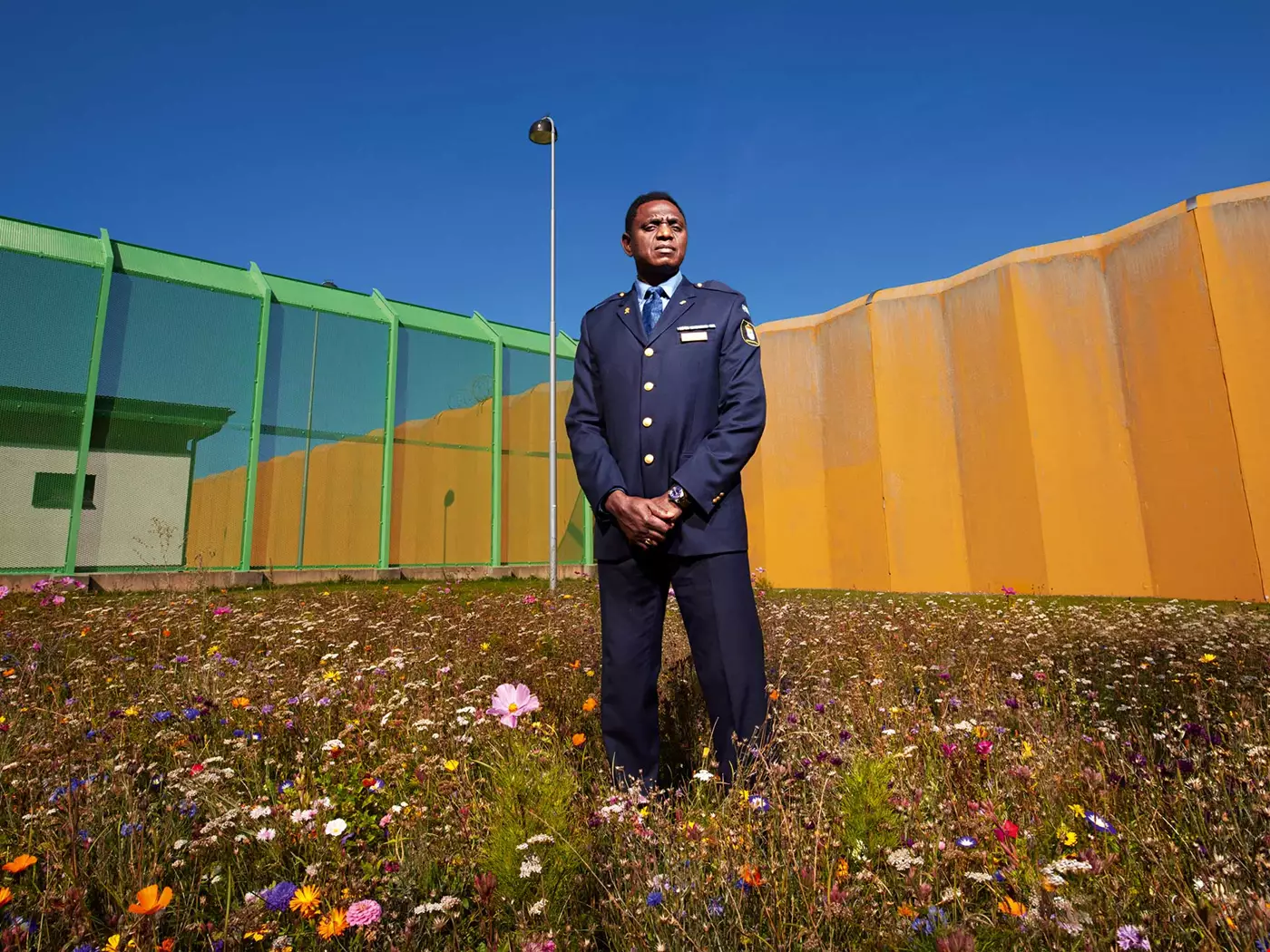 Medelålders man med uniform, står på en blommande sommaräng framför en flera meter hög gul cementmur samt en gallermur som omger ett fängelse