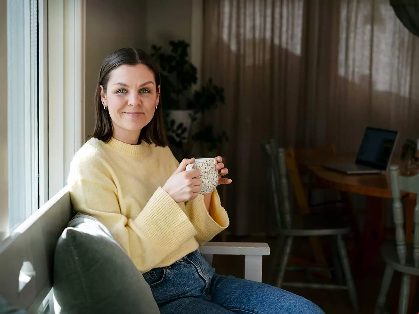 En kvinna med mörkt hår sitter i en grå soffa i en soffa som kan vara på ett kontor, hon ser in kameran och håller en prickig mugg i handen