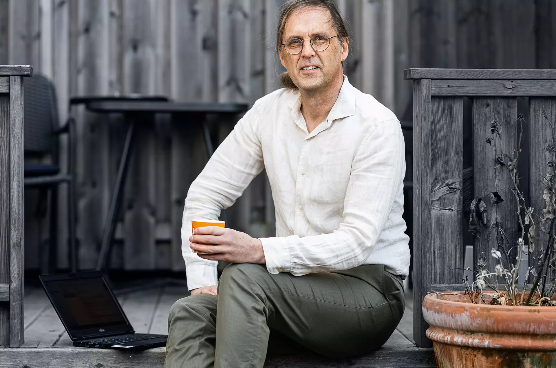 Medelålders man med tonade glasögon, vit skjorta, sitter utomhus vid en silvergrå trävägg. Han håller en röd kaffemugg i handen.